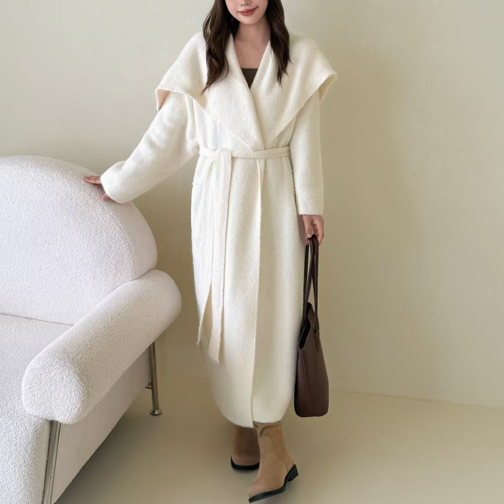 Woman wearing a long white coat holding a brown bag in a minimalistic room.
