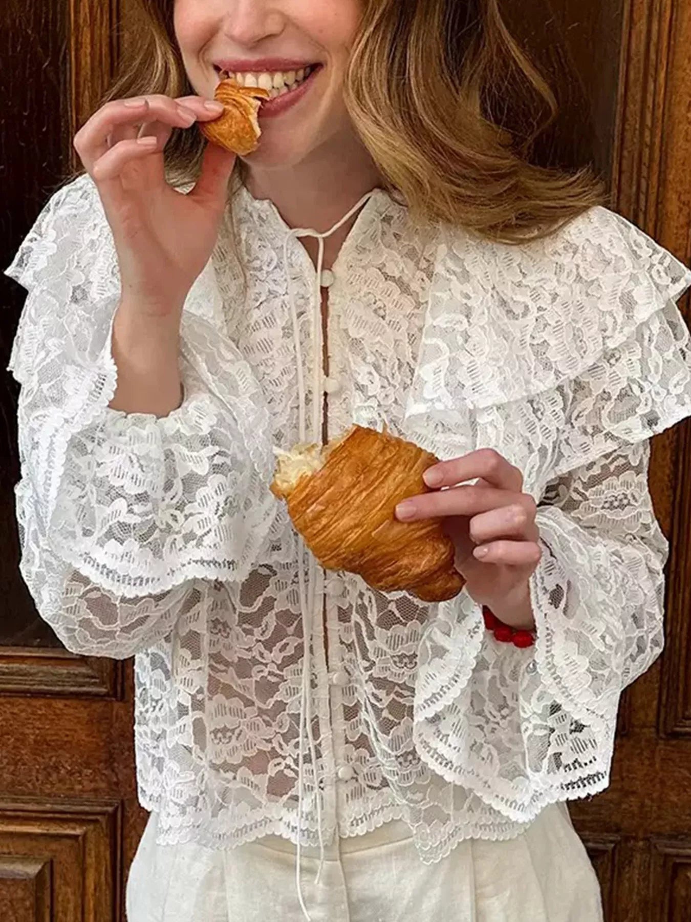Woman in a white lace outfit eating a croissant indoors.