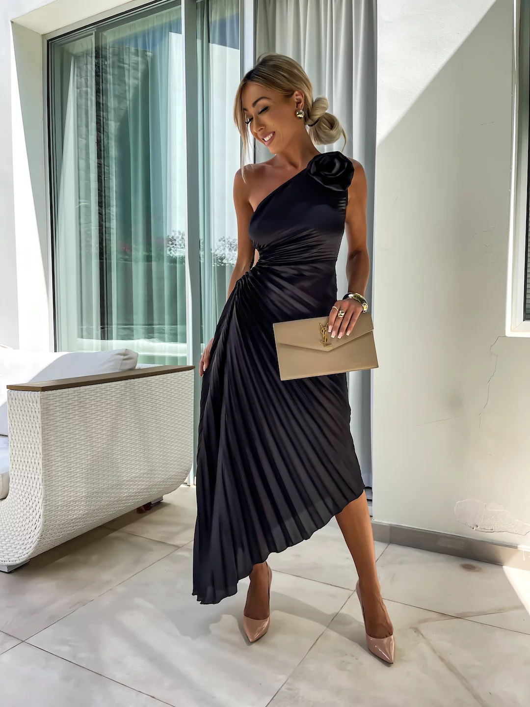Woman in a black pleated dress with a beige clutch in an indoor setting.