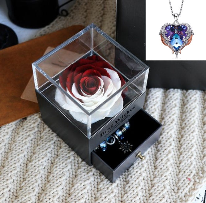 Glass box with a preserved rose and jewelry on a textured surface, with a heart-shaped pendant inset.
