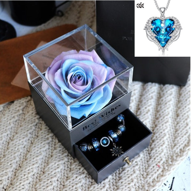 Glass box with a blue and purple rose, bracelet, and necklace on a textured surface.