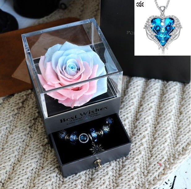 Glass box with preserved pink and blue rose, jewelry set, and blue heart-shaped pendant on a textured surface.