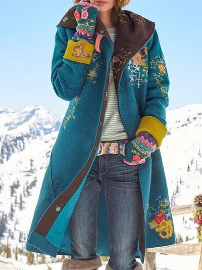 Person wearing a long teal coat with floral embroidery in a snowy mountain setting
