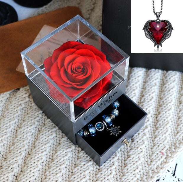 Clear box with a preserved red rose, black jewelry box with necklaces, and heart-shaped pendant on a textured surface.