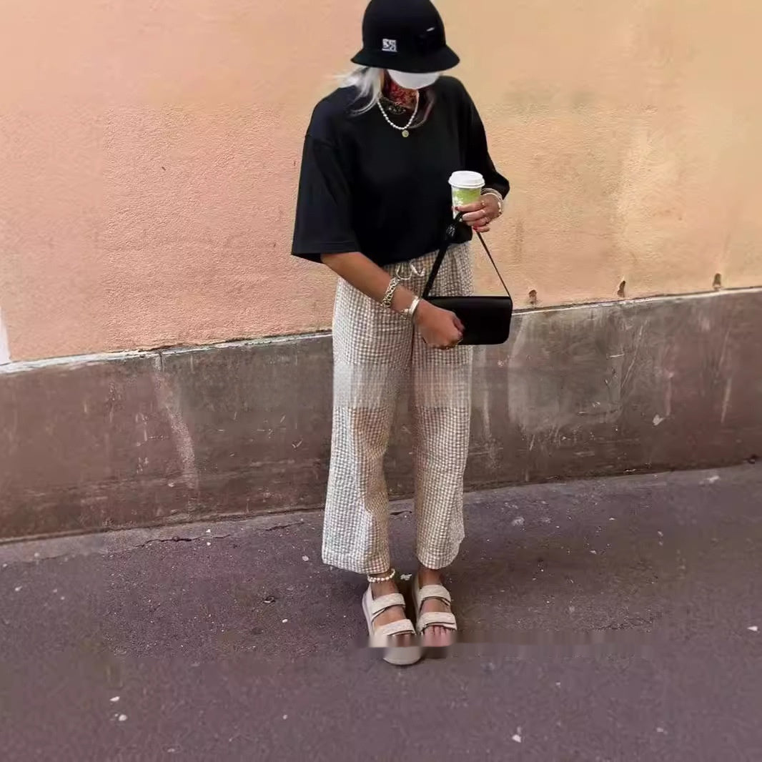 Person wearing a black outfit with checkered pants, holding a coffee cup against a beige wall.