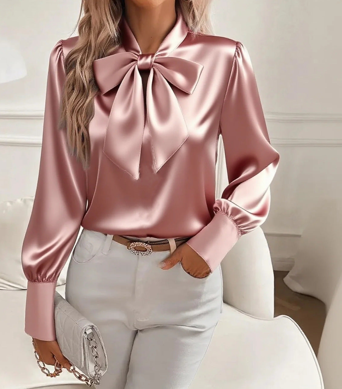 Pink satin blouse with a large bow and white pants in a neutral setting