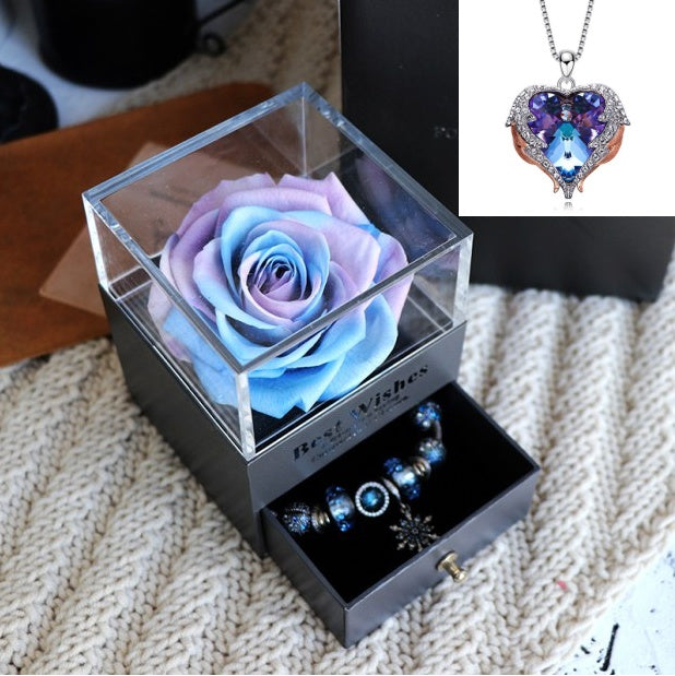 Glass box with a blue and purple rose, jewelry, and a heart-shaped necklace on a textured surface.