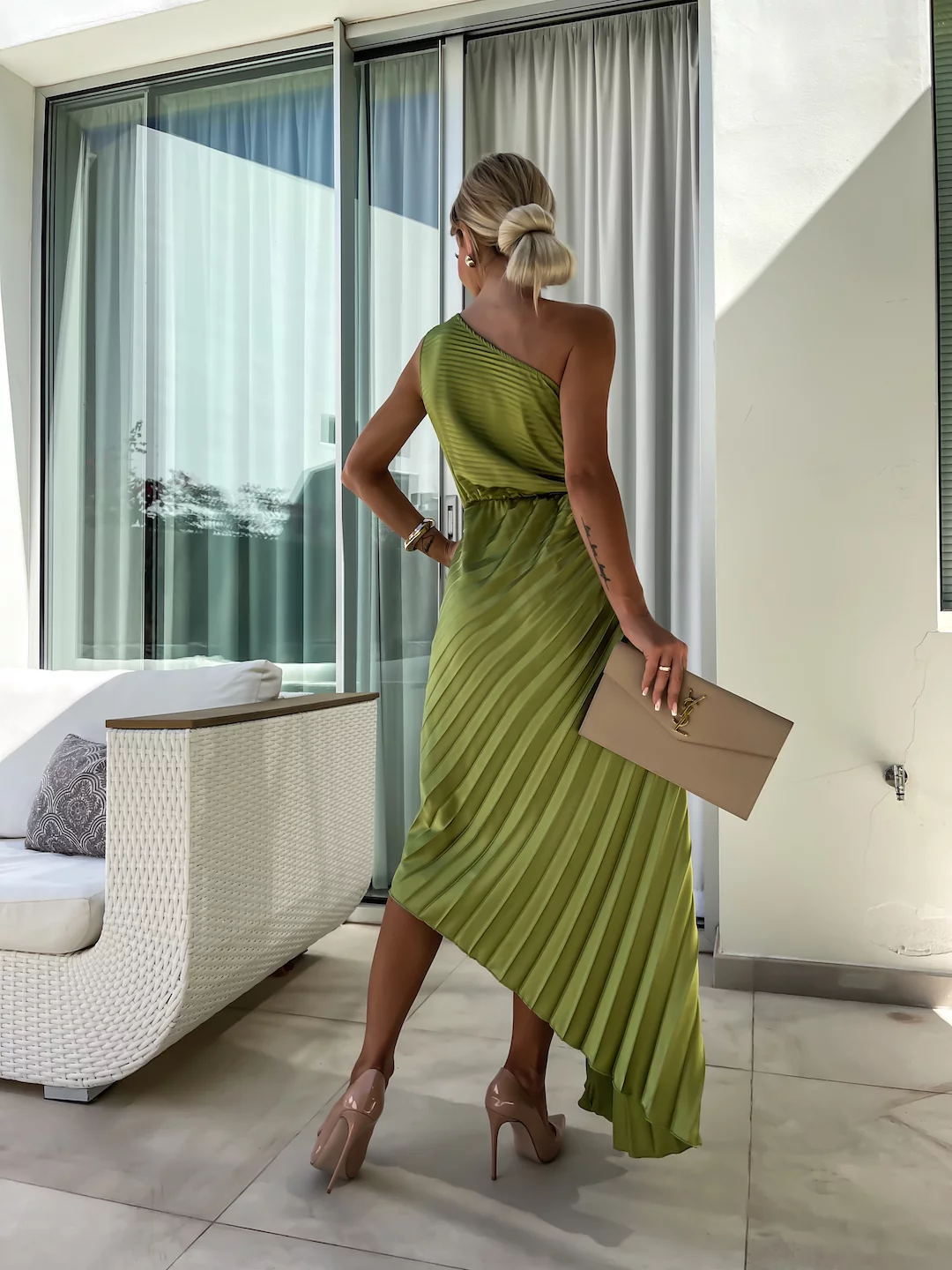 Woman in a green dress holding a beige clutch in an indoor setting with large windows.