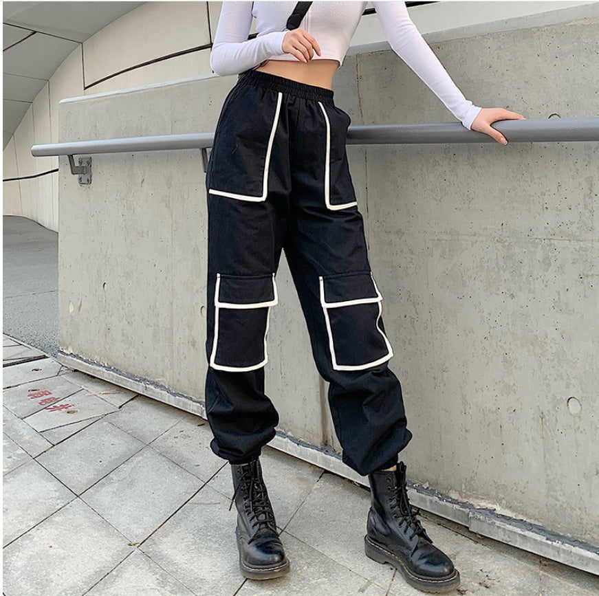 Person wearing black cargo pants with white piping standing against a concrete wall.