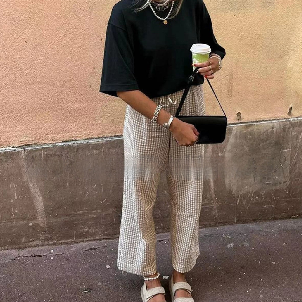 Person wearing a black top and checkered pants, holding a coffee cup and black handbag against a beige wall.