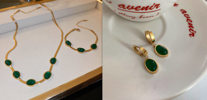 Gold necklace and bracelet with green stones on a white surface, next to gold earrings on a branded coaster.