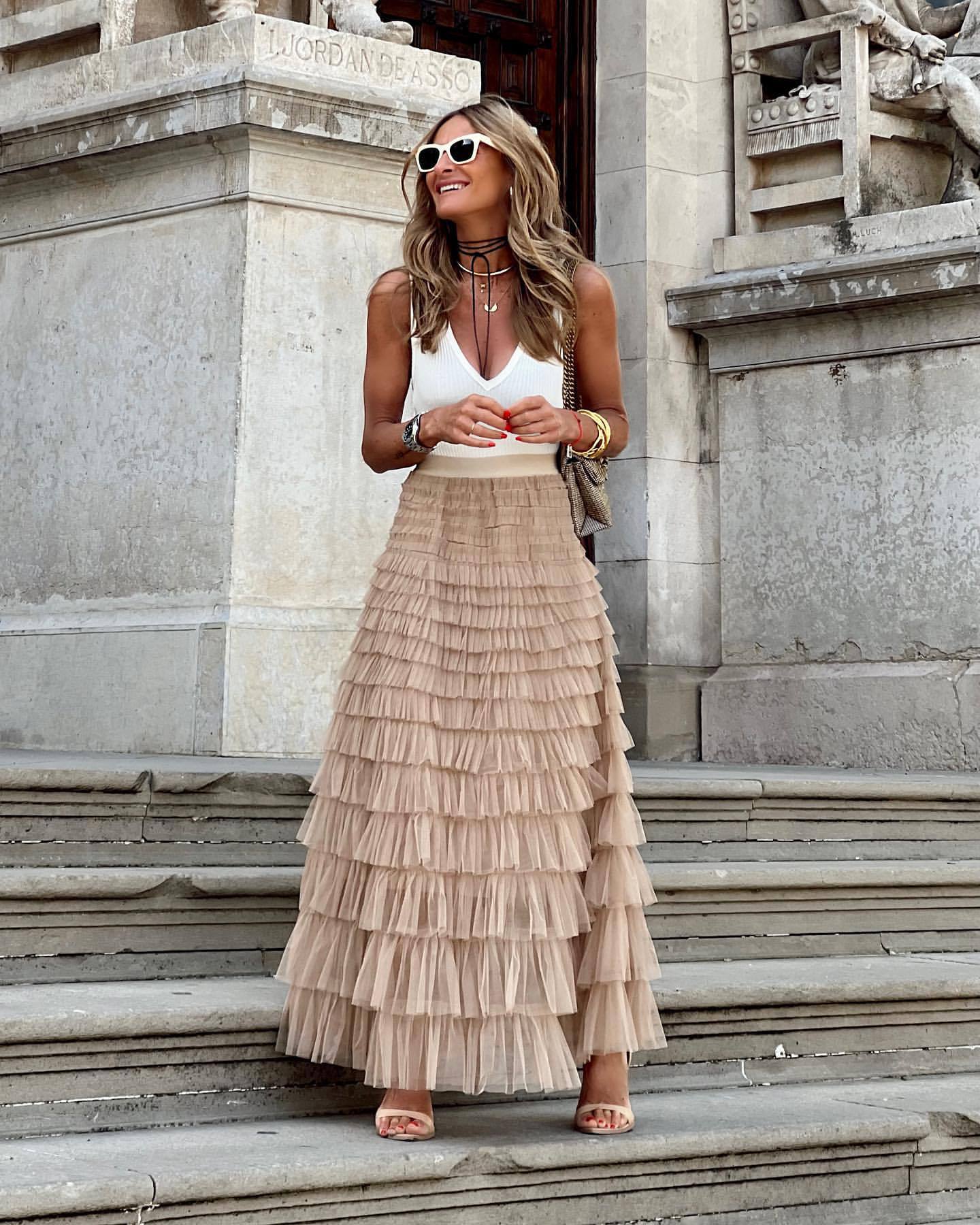 Woman wearing a white top and beige tiered skirt standing on stone steps with classical architecture in the background.