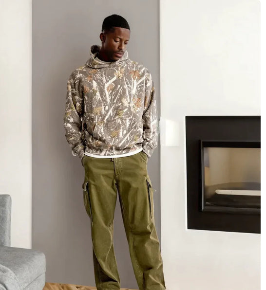 Man wearing a camouflage hoodie and green pants standing in a modern living room.