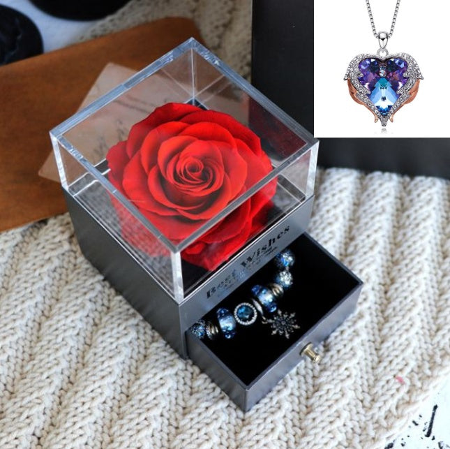Clear box with a red rose, necklace, and earrings on a textured surface.