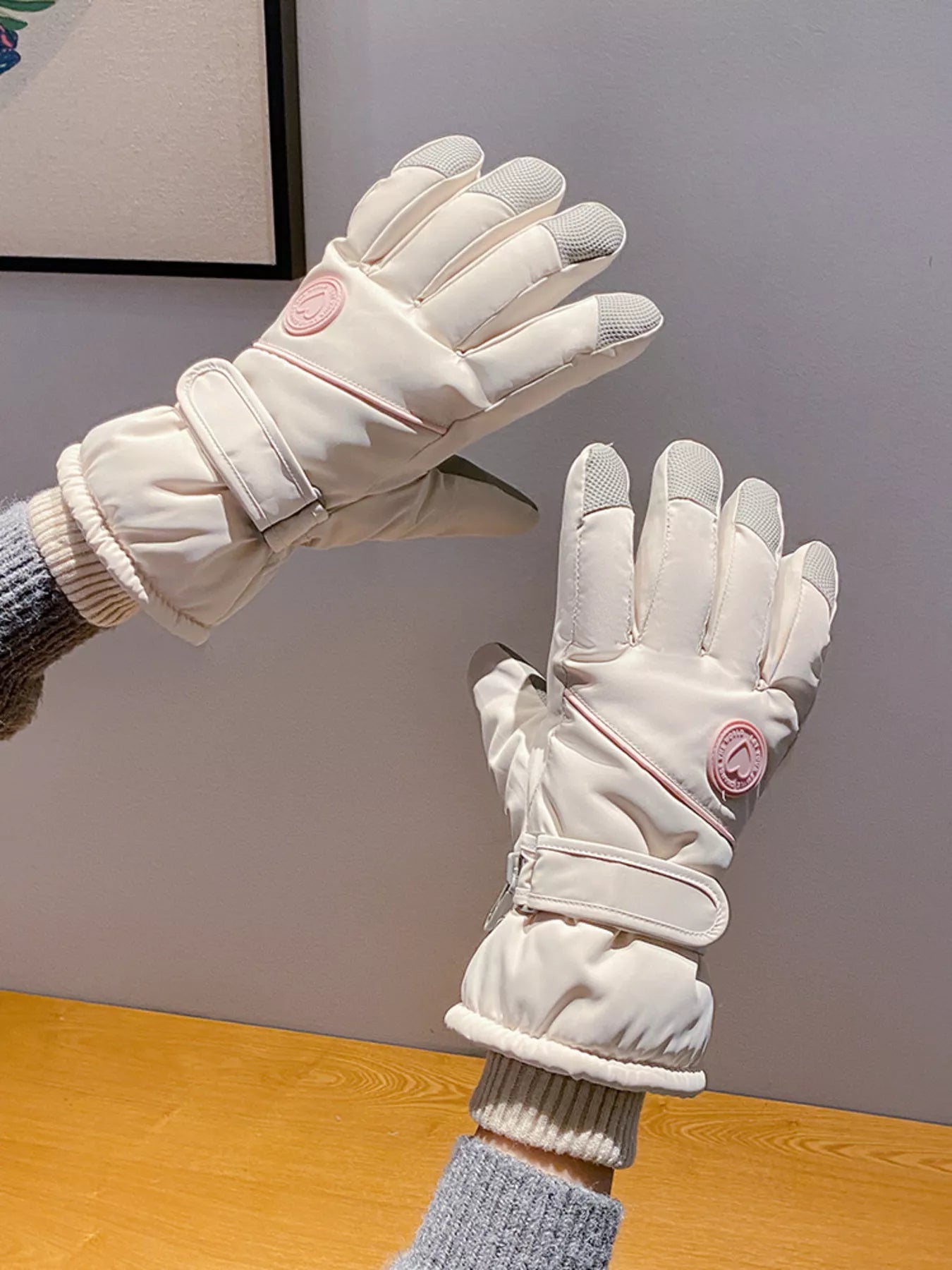 White gloves with a pink logo held by a person wearing gray sleeves on a wooden surface.
