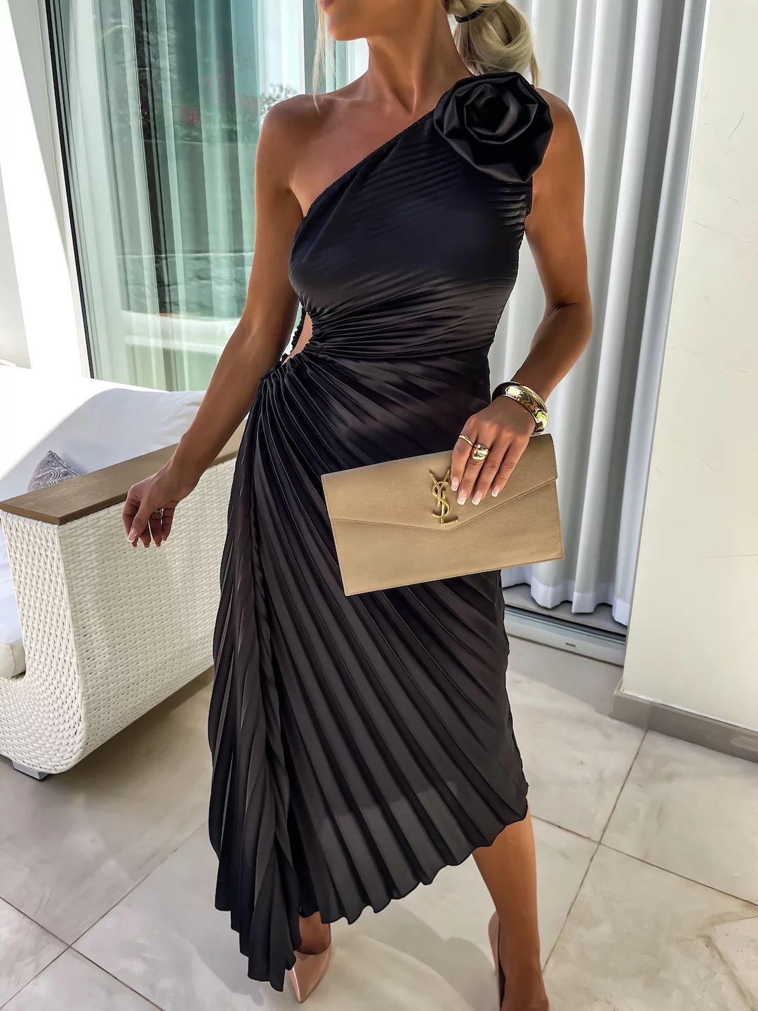 Woman wearing a black one-shoulder dress with a large flower detail, holding a beige clutch.