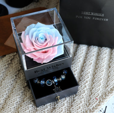Clear box with a pink and blue rose inside, opened to reveal jewelry on a textured surface.