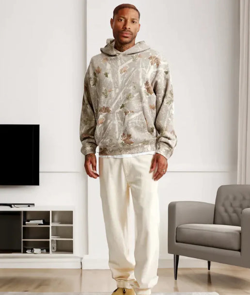 Man wearing a patterned hoodie and white pants in a modern living room.
